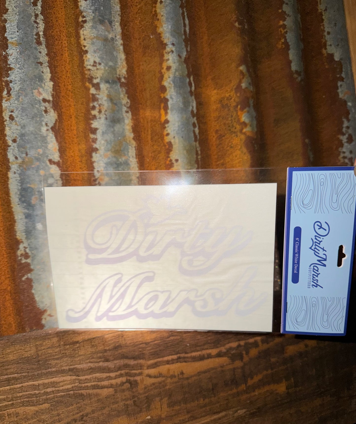 White 6" Dirty Marsh Classic outdoor decal on a clear sheet, next to blue packaging, against a rustic metal and wood background.