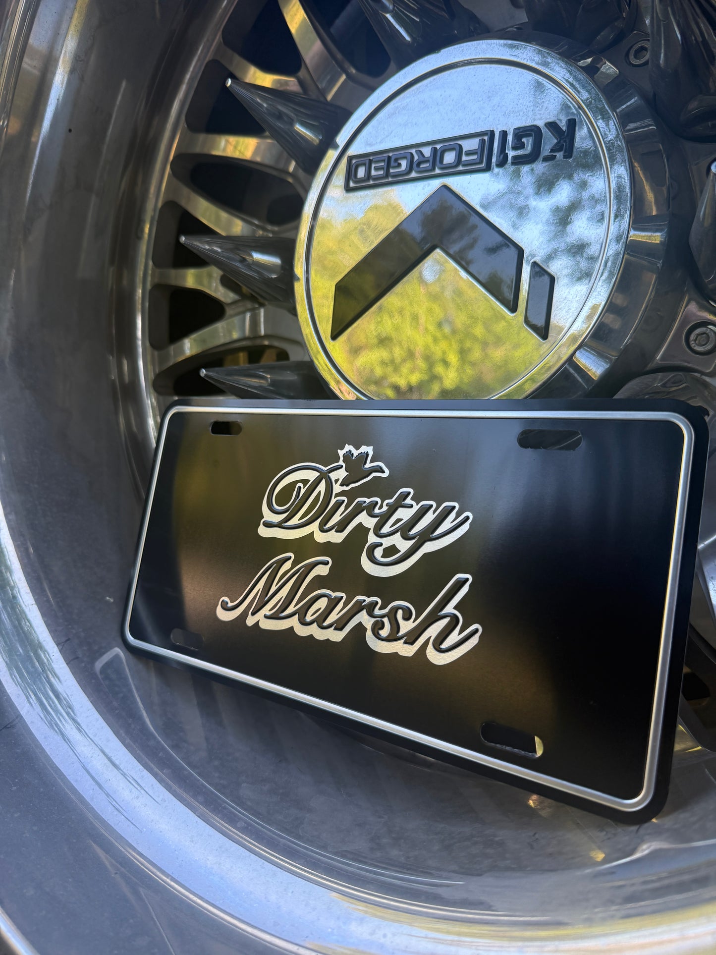 Black aluminum Dirty Marsh license plate with white script logo, leaning on a chrome wheel.