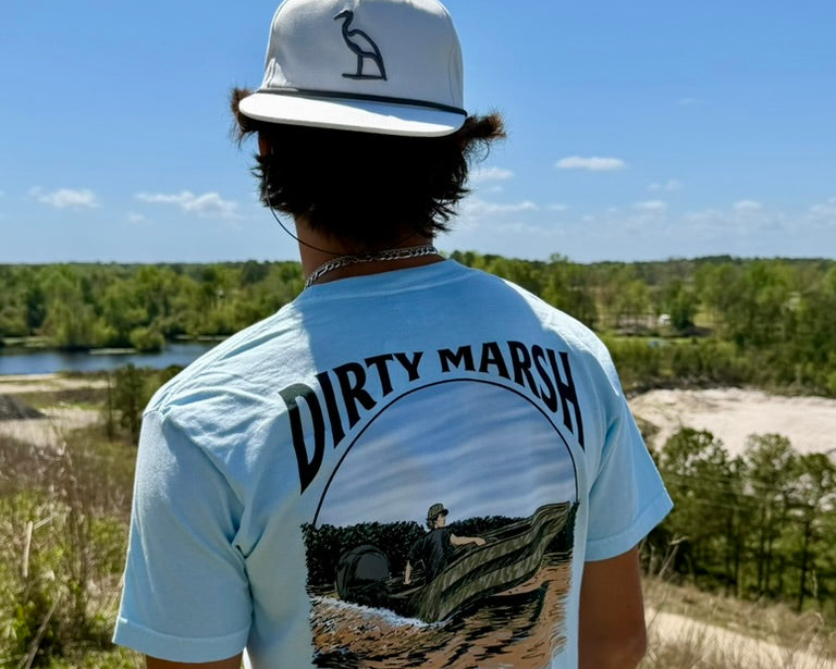 Person wearing a 'DIRTY MARSH' t-shirt and cap, standing in a natural landscape.