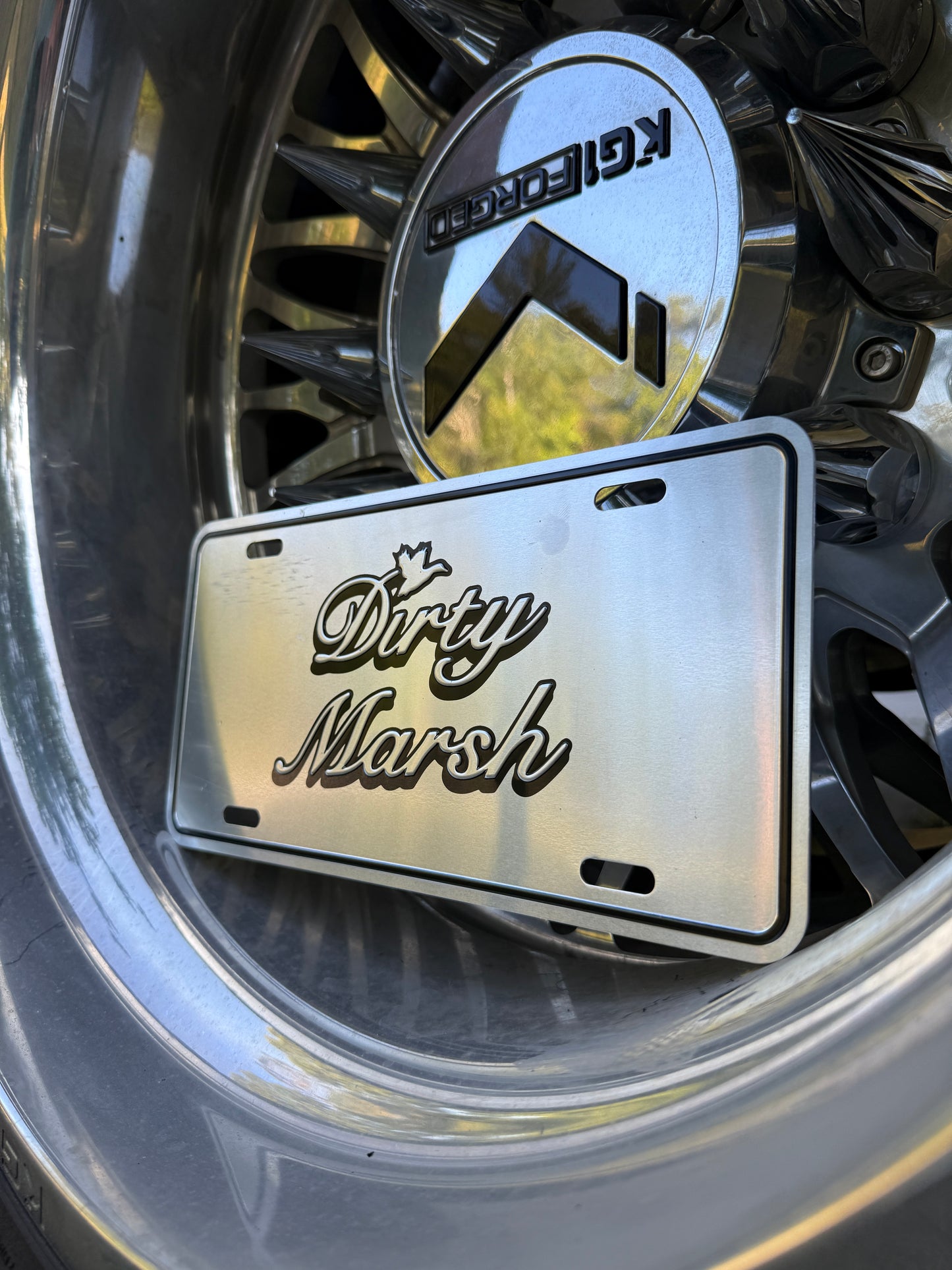 Chrome aluminum license plate with "Dirty Marsh" logo, resting on a shiny chrome wheel.