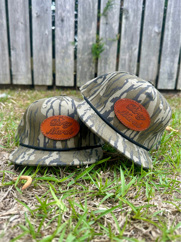 Two Dirty Marsh Youth Classic Leather Patch Hats in Mossy Oak Bottomland Camo, featuring a brown leather patch and black rope detail, resting on grass.