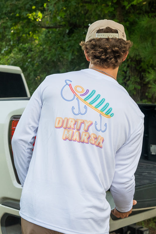 A man viewed from behind wearing a white long-sleeve shirt with a colorful fishing lure graphic and the text 'DIRTY MARSh' on the back, standing next to a white truck outdoors.