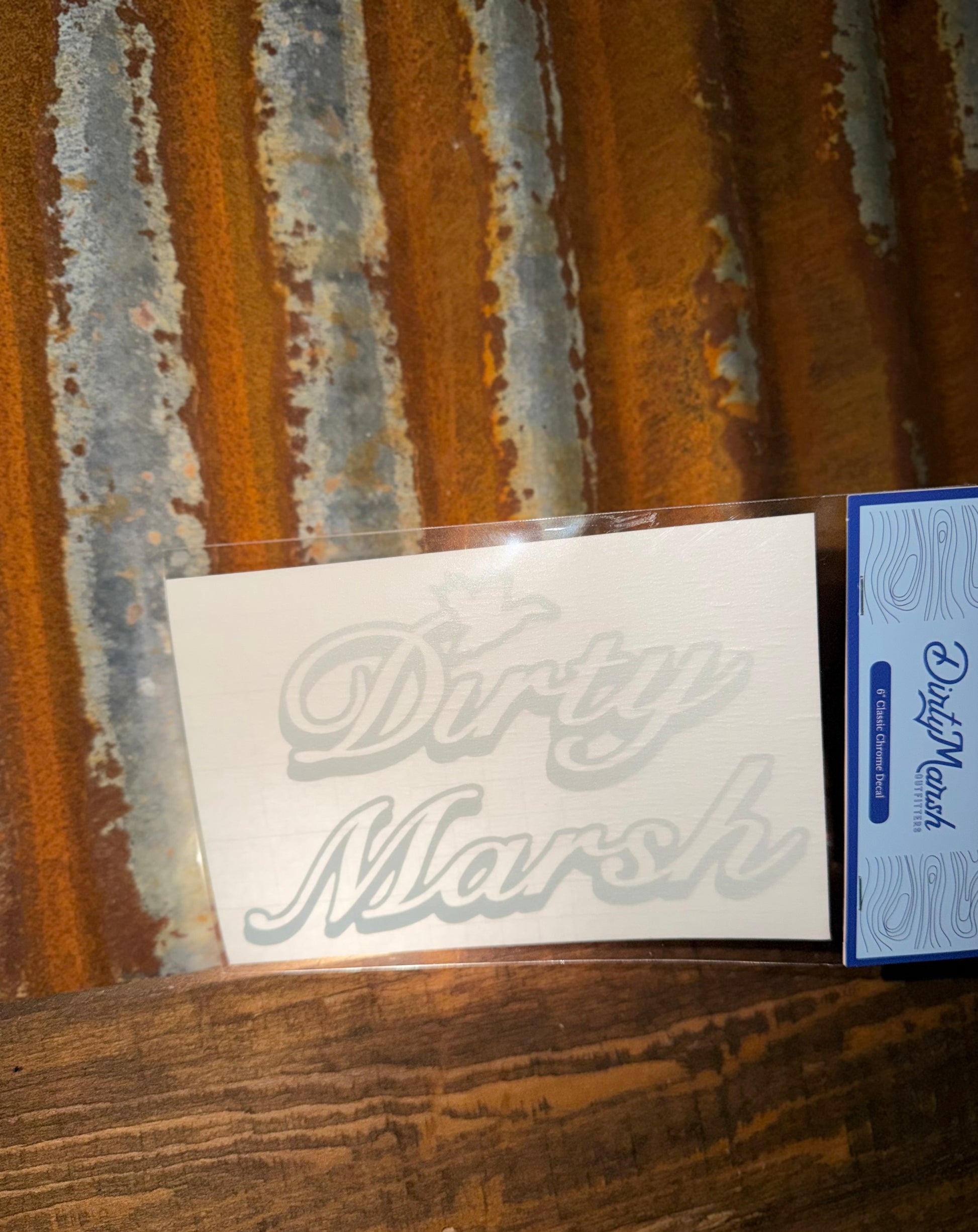 Chrome "Dirty Marsh" 6" decal on clear backing, shown against a rustic metal and wood background.