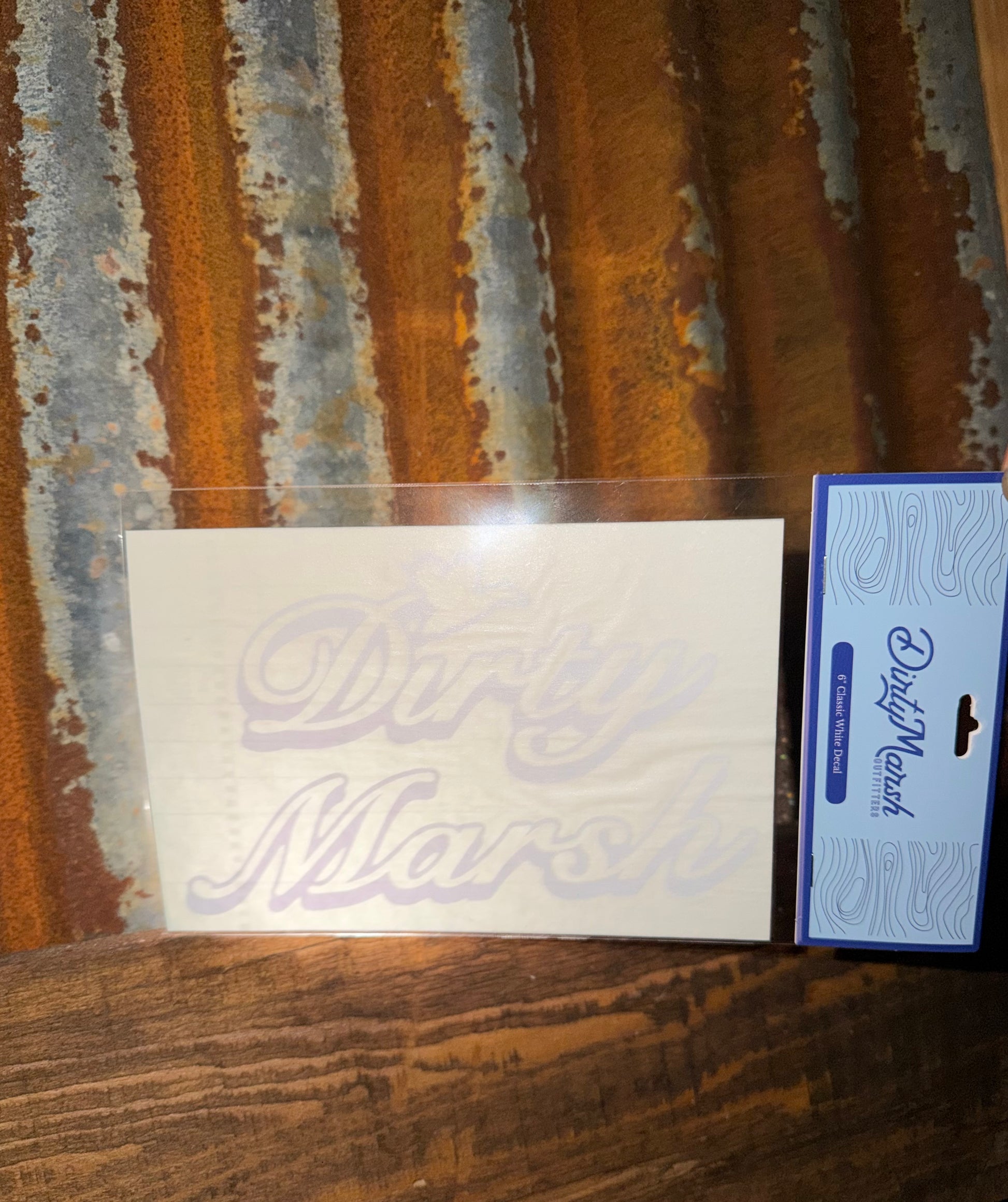 White 6" Dirty Marsh Classic outdoor decal on a clear sheet, next to blue packaging, against a rustic metal and wood background.