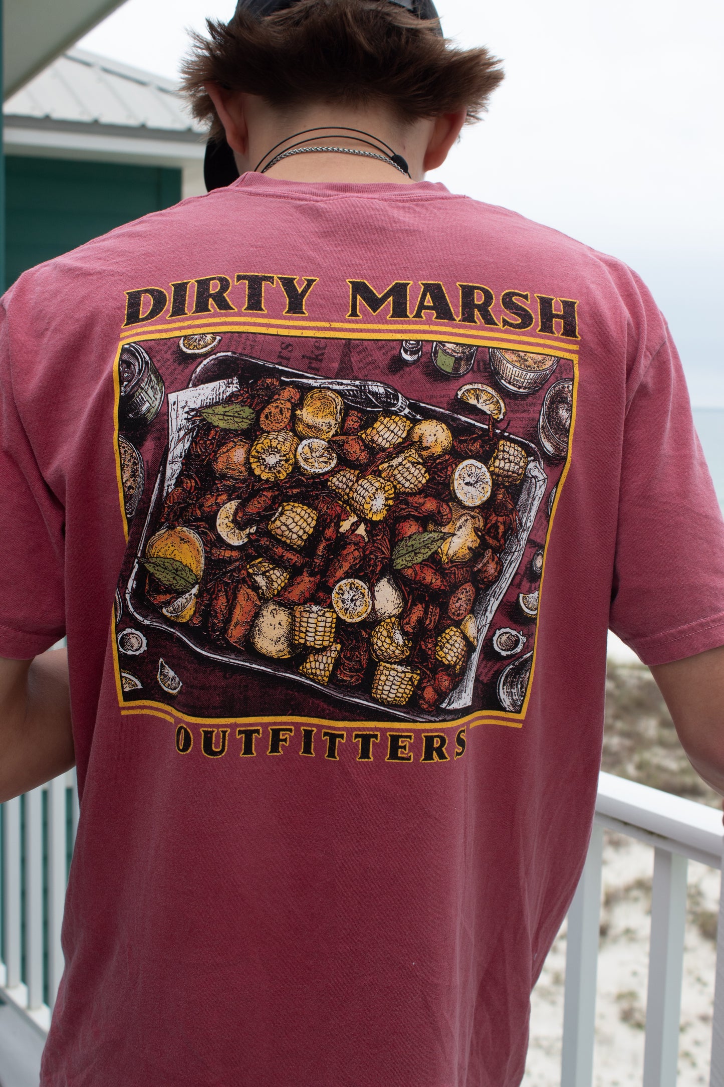 Red t-shirt with a detailed crawfish boil graphic featuring mudbugs, corn, and lemons, "Dirty Marsh Outfitters" text.